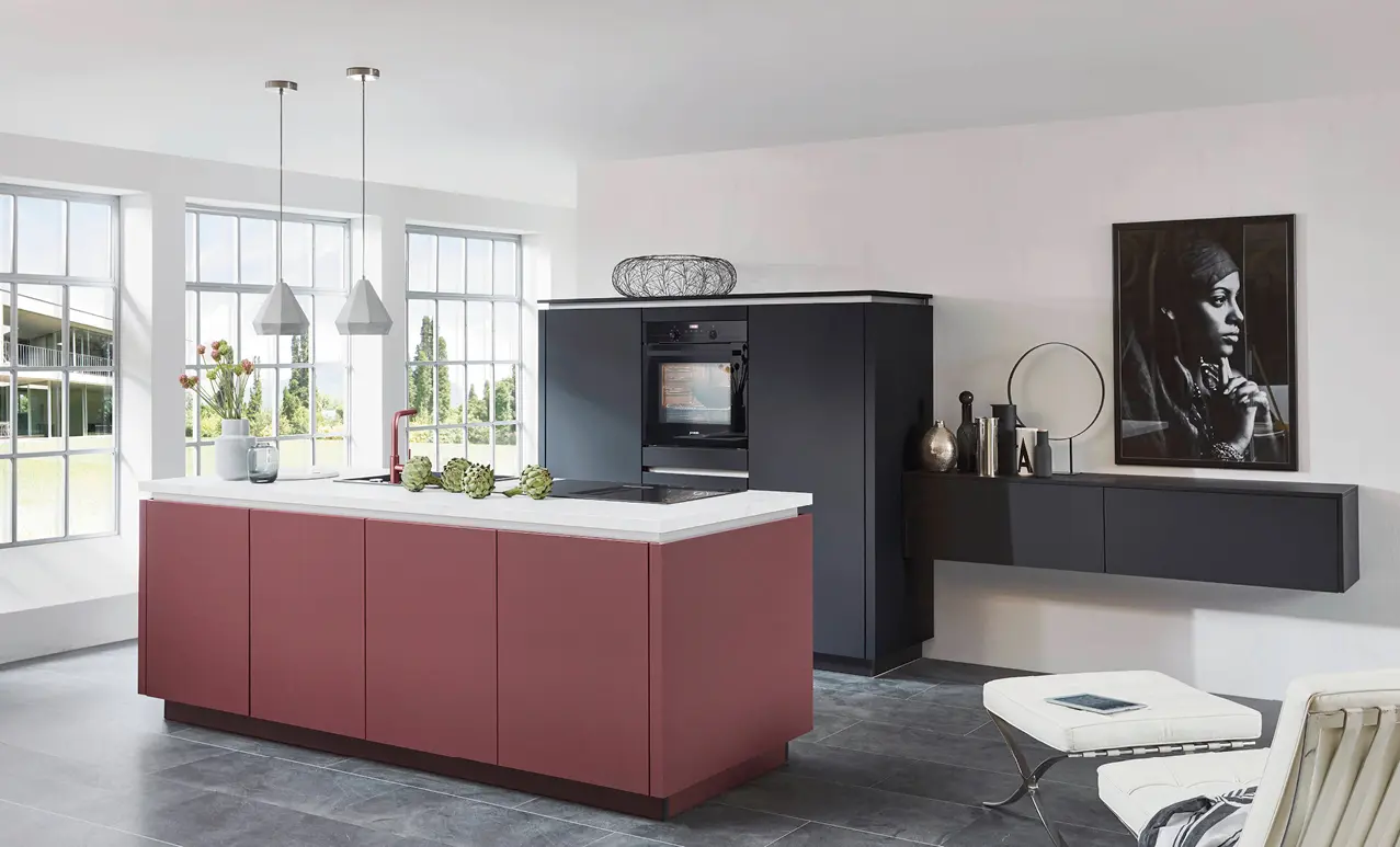 Weinrote Kochinsel mit heller Arbeitsplatte vor hohen Fenstern, dahinter dunkler Hochschrank mit Backofen und wandhängendes Sideboard mit Deko und großem Wandbild.